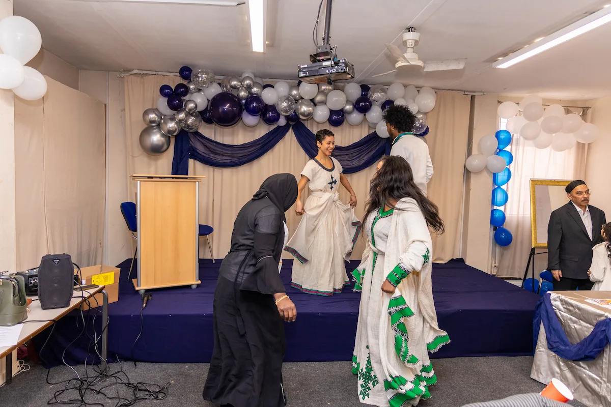 3 Asian women are dancing, one (quite young with short hair) wearing a traditional white dress is on the stage and 2 others, in long dresses (one wearing a headscarf) in front of the stage. The stage is dark purple and the backdrop is a beige curtain with dark purple drapes and purple, silver and white balloons. They are celebrating the achievements of women on the Pathways Without Cobbles project at the ICT Centre in Cheetham Hill Manchester.