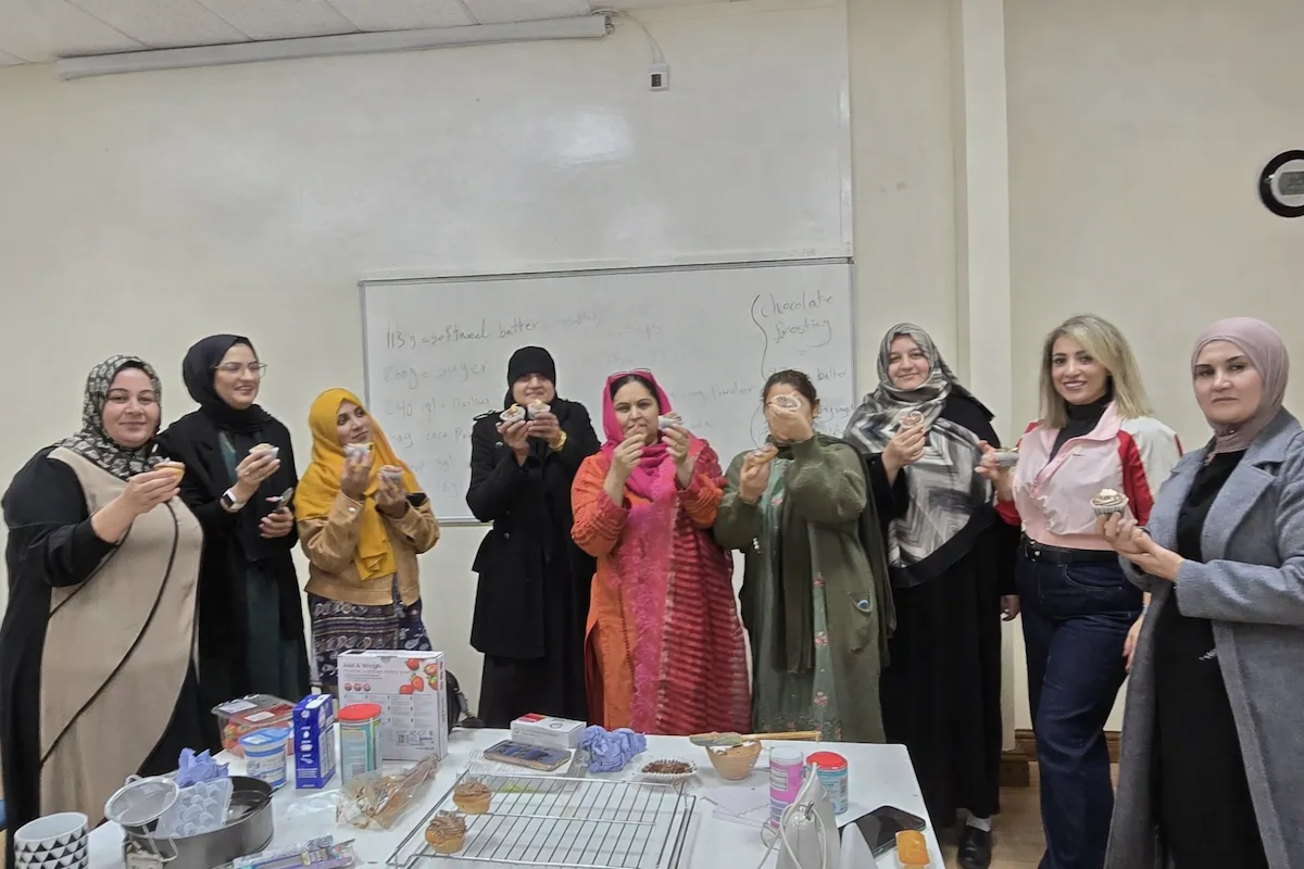 A photograph of 9 women in a line behind a table where they have been baking cakes. The women are mostly South Asian and wearing colourful dresses and scarves. They are happily holding up cakes and smiling. They are in a classroom environment with a whiteboard behind them. They are women who have been part of the Pathways Without Cobbles project and who are interested in running a baking cooperative.