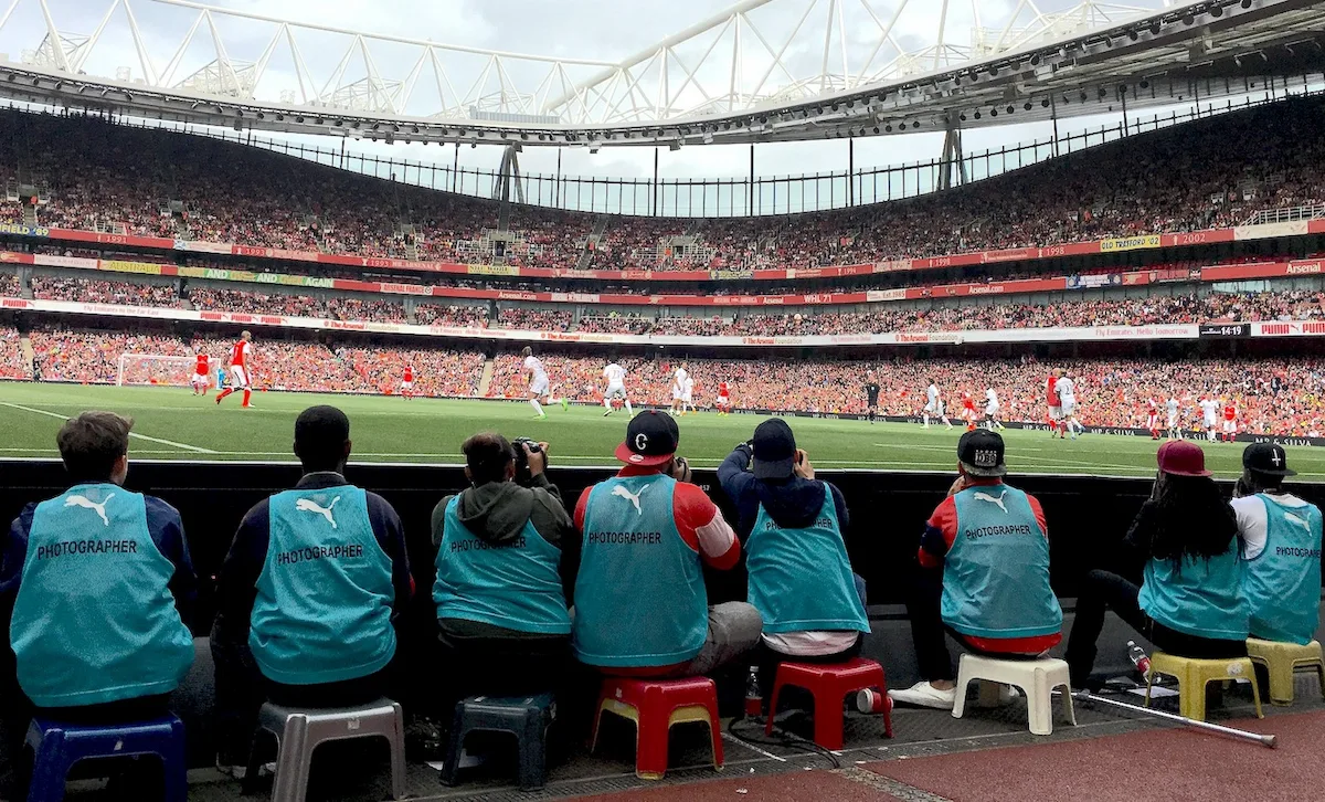 Photo is taken from behind a group of 8 trainee photographers on the sidelines of the football pitch at Arsenal. The young people are from diverse backgrounds and are wearing blue press bibs. They are sitting on low stools right up to the barrier for perfect shots. There is a game on and one team is in red, the other in white. The photo looks out over the pitch and into the stands. It’s a sunny day.